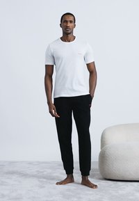 Baltas trumpomis rankovėmis marškinėlis su raudonu logotipu, derinamas su juodomis sportinio stiliaus kelnėmis. Modelis stovi ant šviesiai pilko kilimo šalia smėlio spalvos minkšto sofos.