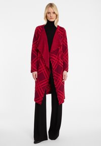 Rot kariertes Strickjacke mit Fransen, aus weichem Material, mit offenem Vorderteil und langen Ärmeln, kombiniert mit schwarzen Hosen.