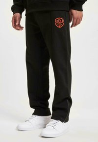 Svart joggebukse med oransje logo, elastisk midje og glidelås detaljer ved anklene. Kombinert med hvite sneakers på en nøytral bakgrunn.