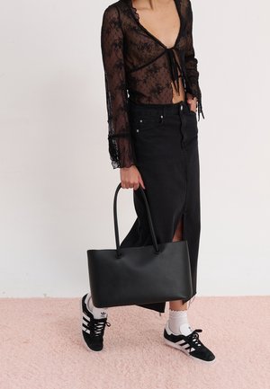 Femme portant un haut en dentelle noire transparente et une jupe midi noire fendue sur le côté, tenant un grand sac cabas noir et portant des baskets Adidas noires avec des chaussettes blanches.
