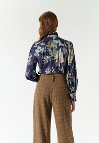 Blouse florale bleu marine à manches longues avec un dos froncé, associée à un pantalon large à carreaux marron avec poches arrière.