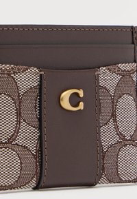 Καφέ πορτοφόλι με υφασμάτινο σχέδιο, λείο δερμάτινο λουράκι και χρυσό μεταλλικό έμβλημα "C" στο κέντρο μπροστά.