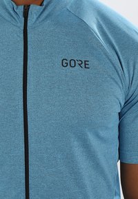 Modré sportovní tričko s krátkým rukávem a zipem, vyrobené z texturované látky; na hrudi je černé logo "GORE".