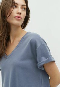 Halenka s výstřihem do V a krátkým rukávem z jemné, světle modré tkaniny s ohrnutými rukávy; hladká textura a volný střih.