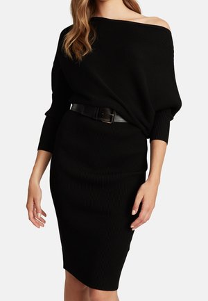 Kobieta w czarnej, prążkowanej sukience dzianinowej z odsłoniętymi ramionami, długimi rękawami i czarnym paskiem w talii.