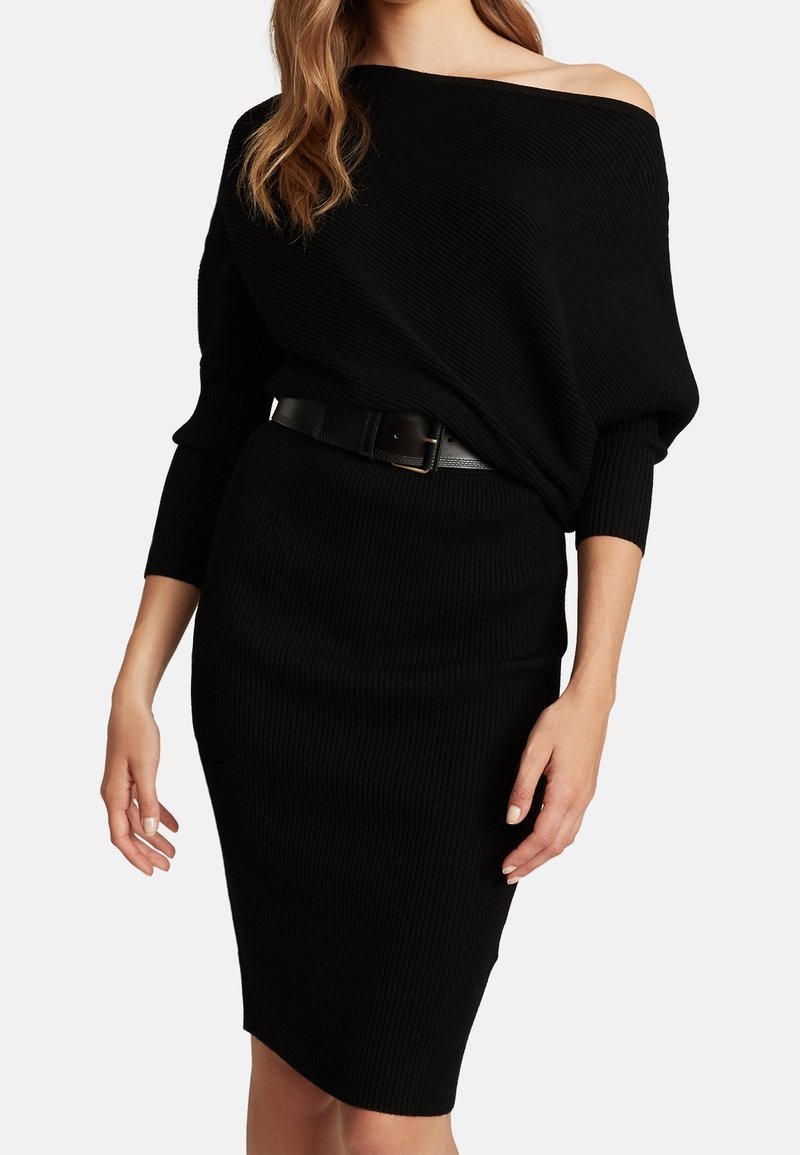 Femme portant une robe noire côtelée à manches longues et épaules dénudées, avec une ceinture noire à la taille.