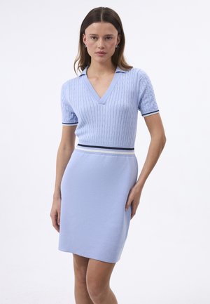 Kobieta ubrana w jasnoniebieską, krótką, dzianą sukienkę z dekoltem w serek, wzorem warkocza oraz paskiem w paski na rękawach i w talii na białym tle.