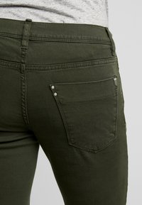 Oliwkowe jeansy o wąskim kroju, z jedną tylną kieszenią i metalowymi nitami. Materiał ma gładką, teksturowaną powierzchnię.