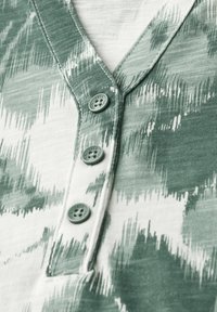 Camisa de patrón verde y blanco con escote en V y tres botones. Tela suave con un aspecto texturizado, con tonos de verde en un diseño de olas.