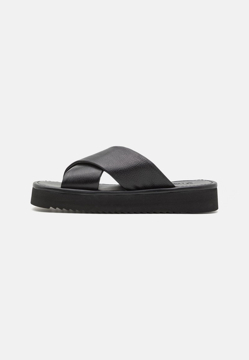 Mule noire avec larges brides croisées en cuir et semelle épaisse texturée sur fond gris clair uni.