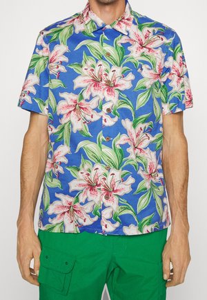 Kurzärmeliges Hemd mit Knöpfen und großem rosa-weißem Blumenmuster auf blauem Hintergrund, getragen mit grünen Cargo-Shorts.