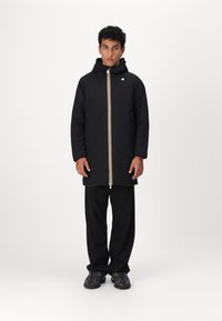 Veste à capuche noire avec accent jaune sur la fermeture éclair et un logo. Associée à un pantalon noir ample et des chaussures de sport sombres. Texture de tissu lisse.
