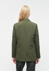 Olivovo zelený blazer so strihom na mieru, jednopremeňovaným dizajnom, klopovými západkami a stredným rozparkom vzadu. Vyrobený z hladkej tkaniny.