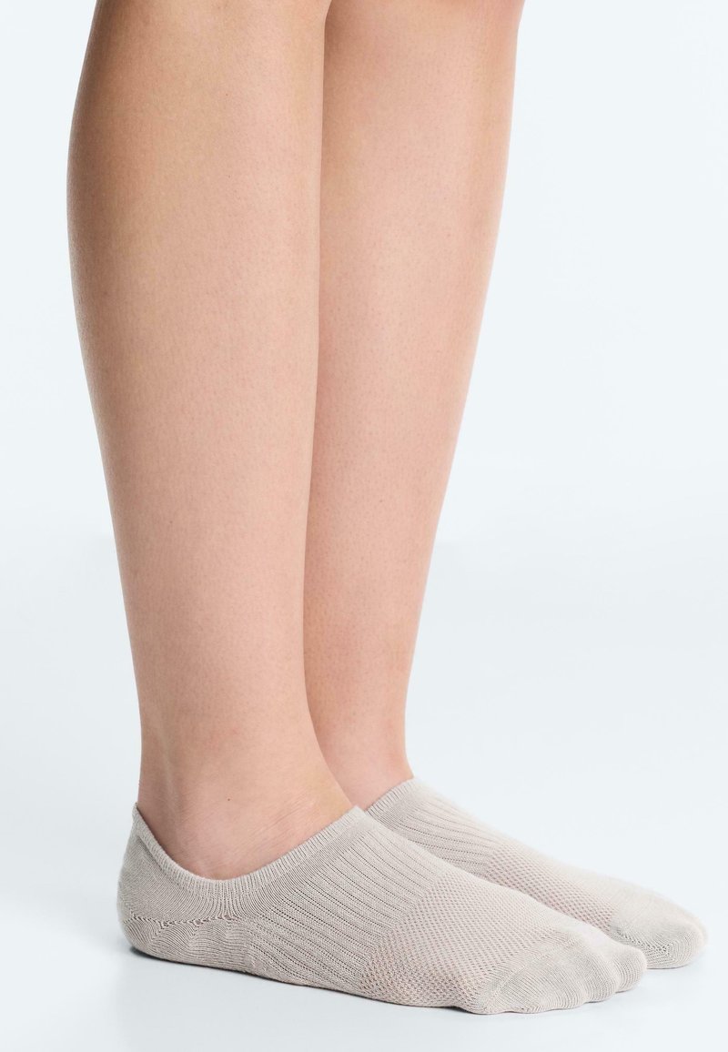 Untere Beine, die hellbeige, nicht sichtbare Socken tragen, stehen auf einer glatten weißen Oberfläche vor einem einfarbigen hellen Hintergrund.