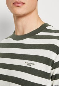 Primer plano de una persona que lleva una camiseta de rayas verdes y blancas con el texto "Marc O'Polo DENIM" impreso en el pecho.