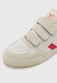 Baltos sportbačiai su raudonais akcentais, iš zomšos ir odos medžiagų, su Velcro lipdukais, perforuota pirštų dalimi ir tekstūruotu padu.