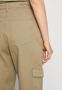 Khaki cargo kalhoty s rovnějším střihem, mající zadní kapsu, viditelné švy a malé logo na pase. Hladká textura.