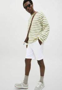 Camisa de manga larga a rayas en verde y blanco, pantalones cortos blancos, zapatillas grises y gafas de sol. La camisa presenta un ajuste relajado y rayas horizontales.