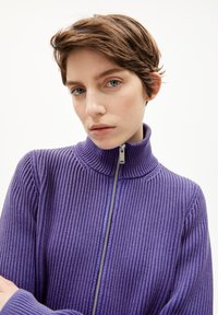 Lila gestrickter Pullover mit hohem Kragen und durchgehendem Reißverschluss, mit geripptem Material und langen Ärmeln. Silberne Reißverschlussbeschläge.