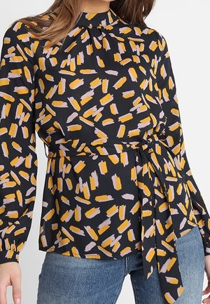 Donna che indossa una camicetta nera con motivo a pennellate arancioni e rosa chiaro, maniche lunghe, collo alto e cintura annodata in vita, abbinata a jeans blu.