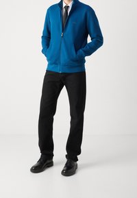 Giacca blu con zip, colletto alto, polsini a costine e tasche laterali, abbinata a pantaloni neri e scarpe nere. Logo semplice sul petto.
