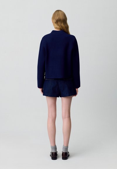 Maglione corto in misto lana blu navy abbinato a jeans corti scuri. Modello indossa calze grigie e scarpe marroni, visto da dietro su uno sfondo chiaro.