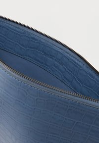 Sinine krokodillinahast reljeefne nahkkott tumeda metallist luku ja siledapoolse sisuga, mille serval on diskreetne kaubamärgi reljeef.