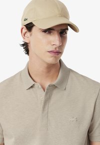 Beige pikétröja med en texturerad tygkvalitet och en kort placket, med en diskret broderad logotyp på vänster bröst. Ljussandy keps kompletterar stilen.