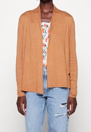 Person trägt eine hellbraune, offene Strickjacke, ein florales Oberteil und hellblaue, zerrissene Jeans, zu sehen vom Hals bis zur Hüfte.