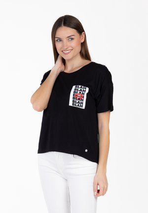Femme souriante aux longs cheveux bruns portant un T-shirt noir avec le texte répété "BLAH" et un graphique de lèvres rouges, associé à un pantalon blanc.