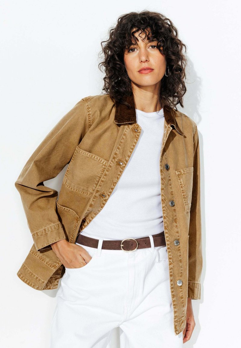 Femme aux cheveux bouclés portant une veste beige sur une chemise blanche et un pantalon blanc, se tenant avec une main dans la poche contre un fond blanc.