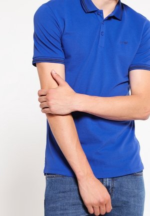 Henkilö, joka pukeutuneena siniseen poolopaitaan ja farkkuihin, pitää vasenta kättään oikealla kädellään yksiväristä taustaa vasten.