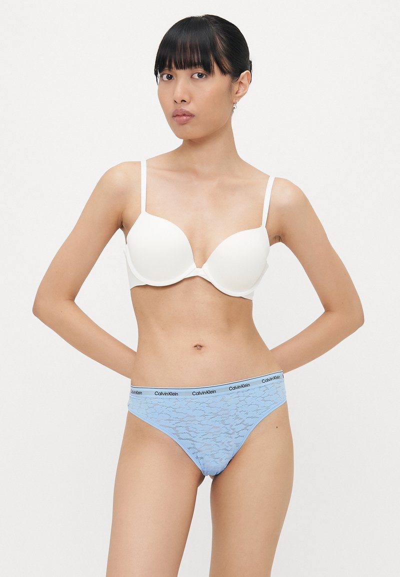 Witte bh met voorgevormde cups en dunne bandjes; blauwe kanten string met Calvin Klein-logo op de tailleband; gladde texturen en een aansluitend design.