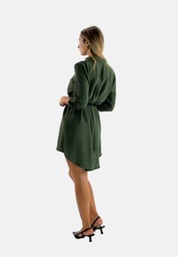 Groene jurk met ceintuur, lange mouwen, een getailleerde taille en een gelaagde zoom; gecombineerd met zwarte sandalen met bandjes. Gladde textuur, ontspannen pasvorm.