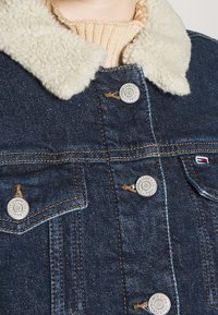Denim jacket with cream sherpa collar, metal buttons labeled "Tommy Jeans New York," and tan knit sweater visible underneath.