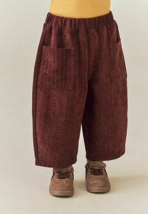 Benen met losse, wijd uitlopende bordeaux ribfluwelen broeken met voorzakken en bruine sneakers, tegen een effen lichte achtergrond.