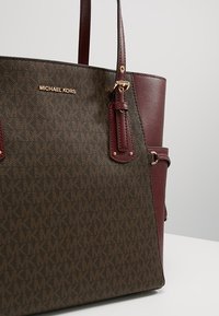 Sac fourre-tout marron Michael Kors avec le motif répété du logo MK et des poignées en cuir rouge foncé ainsi que des panneaux latéraux avec ferrures dorées.