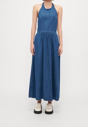 Ärmelloses blaues Denim-Maxi-Kleid mit Neckholder, geraffter Taille, getragen mit weißen klobigen Loafers vor einfarbigem Hintergrund.