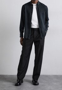 Camicia button-up scura sopra una t-shirt bianca, abbinata a pantaloni neri a righe sottili e scarpe nere lucide, in piedi su uno sfondo neutro.
