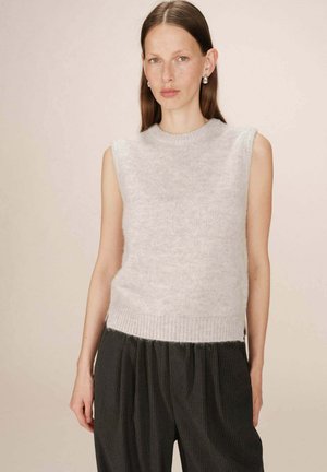 Femme portant un top en tricot léger sans manches gris clair et un pantalon noir à rayures, se tenant devant un fond beige uni.