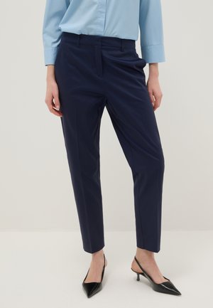 Mujer con pantalones de vestir azul marino ajustados, camisa azul claro de manga larga y zapatos de tacón bajo negros de punta puntiaguda, de pie sobre un suelo blanco.