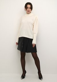 Pull en maille crémeux avec col roulé et manches bouffantes, associé à une jupe A-line en simili cuir noire. Le mannequin porte également des escarpins noirs.