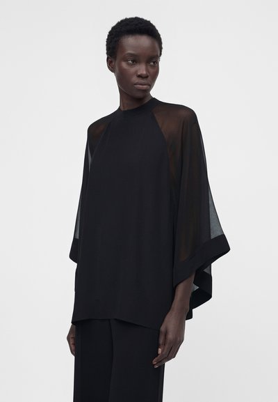 Γυναίκα που φοράει άνετο μαύρο μπλουζάκι με διάφανα, φαρδιά μανίκια, στέκεται μπροστά σε ένα μονόχρωμο ανοιχτό φόντο, κοιτάζοντας ελαφρώς στο πλάι.