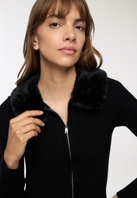 Kobieta w czarnym swetrze z prążkowanej tkaniny z zamkiem błyskawicznym i czarnym kołnierzem z sztucznego futra, trzymająca kołnierz jedną ręką na jednolitym tle.