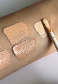 Quatre teintes de fond de teint liquide appliquées sur la peau d'un bras, allant du beige clair au beige moyen, avec un pinceau appliquant la teinte la plus claire.