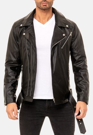 Homme portant une veste de motard en cuir noire avec des fermetures éclair argentées et un t-shirt blanc à col en V, debout, les mains près de la taille.