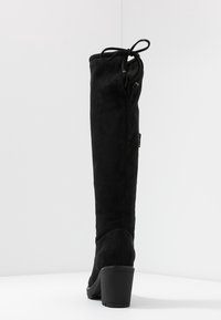 Botte en suède noire, haute jusqu'au genou, avec un talon carré, dotée d'un détail noué en haut et d'une texture lisse. Fermeture éclair sur le côté pour un accès facile.