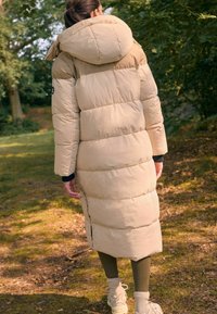 Beige lang pufferjakke med hætte, delt op i vandrette sektioner, har en fuld lynlås og sideslidser for bevægelsesfrihed, båret over grønne leggings.