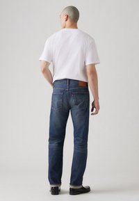 Jeans de mezclilla azul con un corte recto, que presentan un parche trasero de cuero marrón, bolsillos traseros y un ligero desgaste. Combinados con una camiseta blanca.
