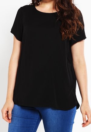 Donna che indossa una maglietta nera a maniche corte e jeans blu, in piedi davanti a uno sfondo bianco.